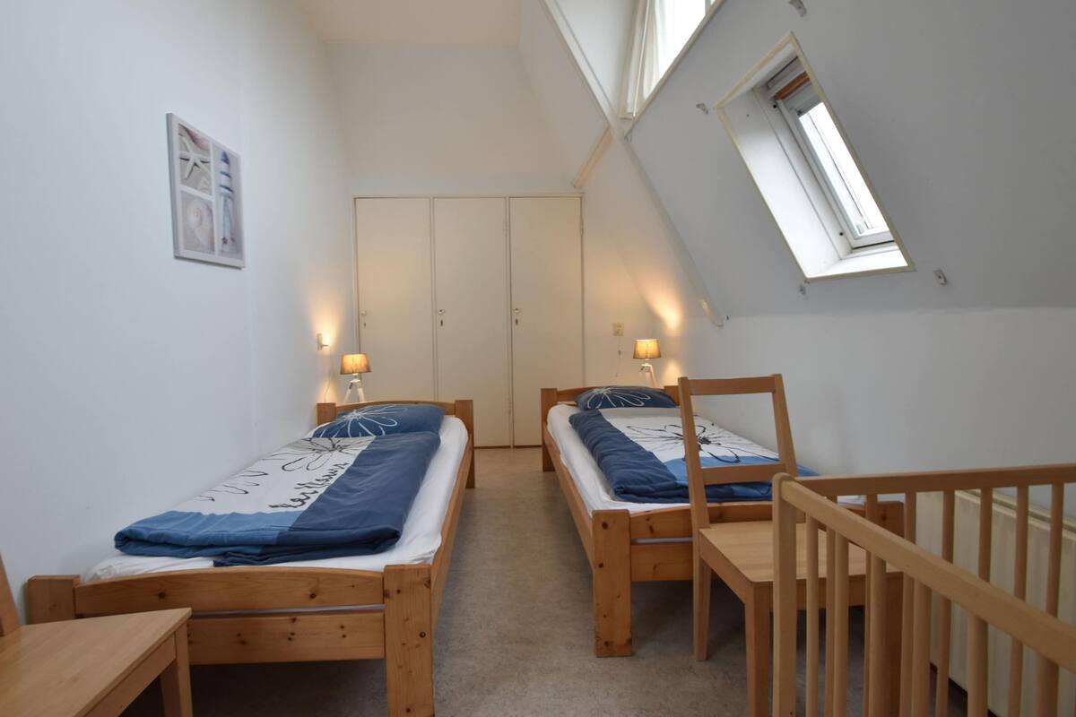 Una habitación pequeña y bien iluminada con dos camas de madera, un escritorio y una silla. Hay una ventana que deja entrar luz natural y una puerta que conduce a otra habitación.