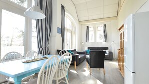 Een lichte, zonnige kamer met een eettafel, stoelen, een bank en een koelkast.