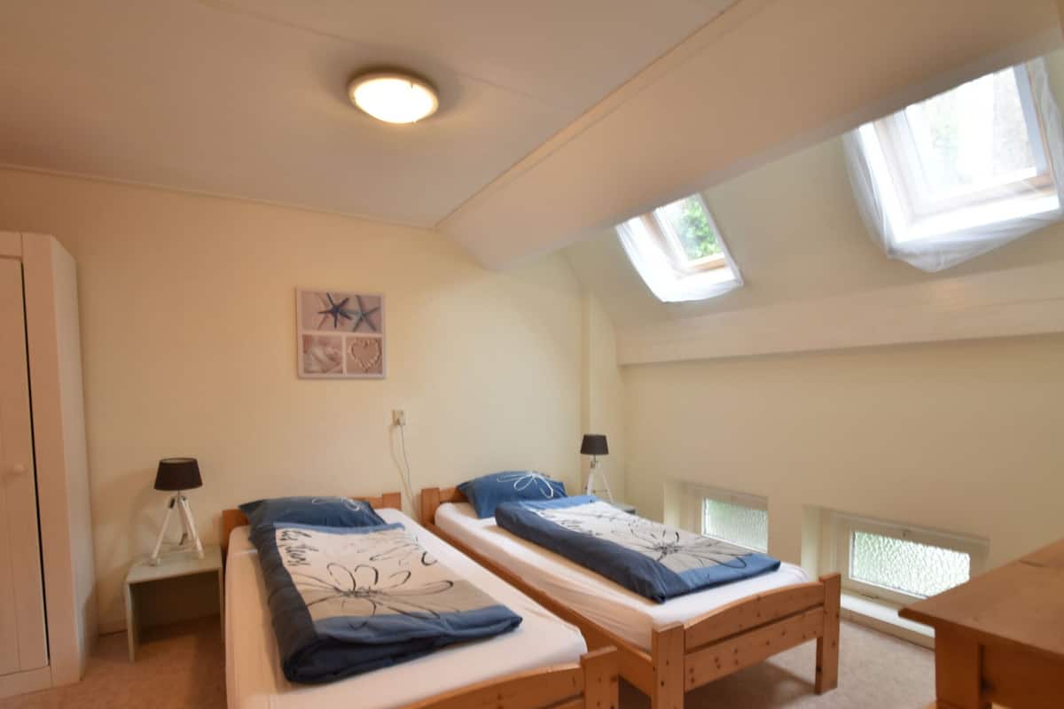 Un dormitorio con dos camas de madera, un escritorio, una mesita, una lámpara y una luz en el techo.
