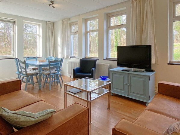A living room with a television, a dining table with chairs, a sofa, and large windows with curtains.