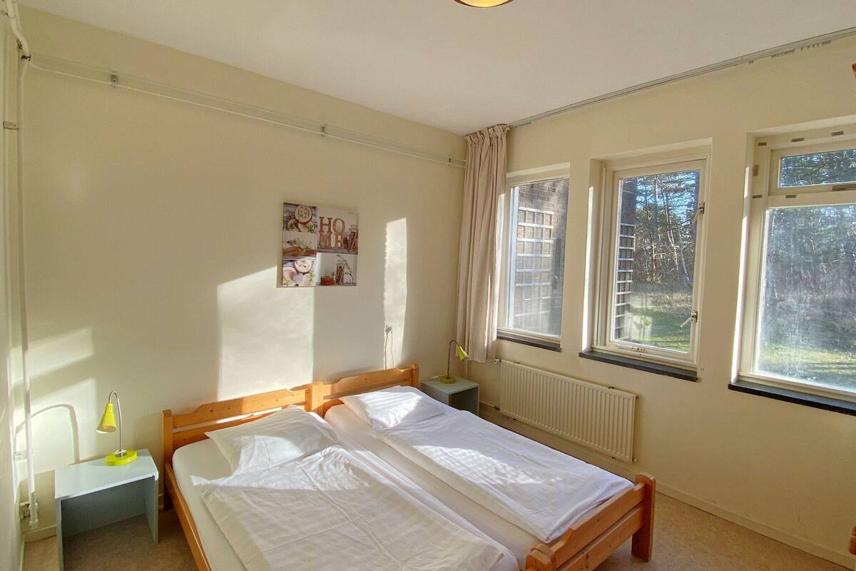 Un dormitorio con dos camas individuales, una mesita de noche con lámpara, un radiador y una ventana con vistas a árboles.