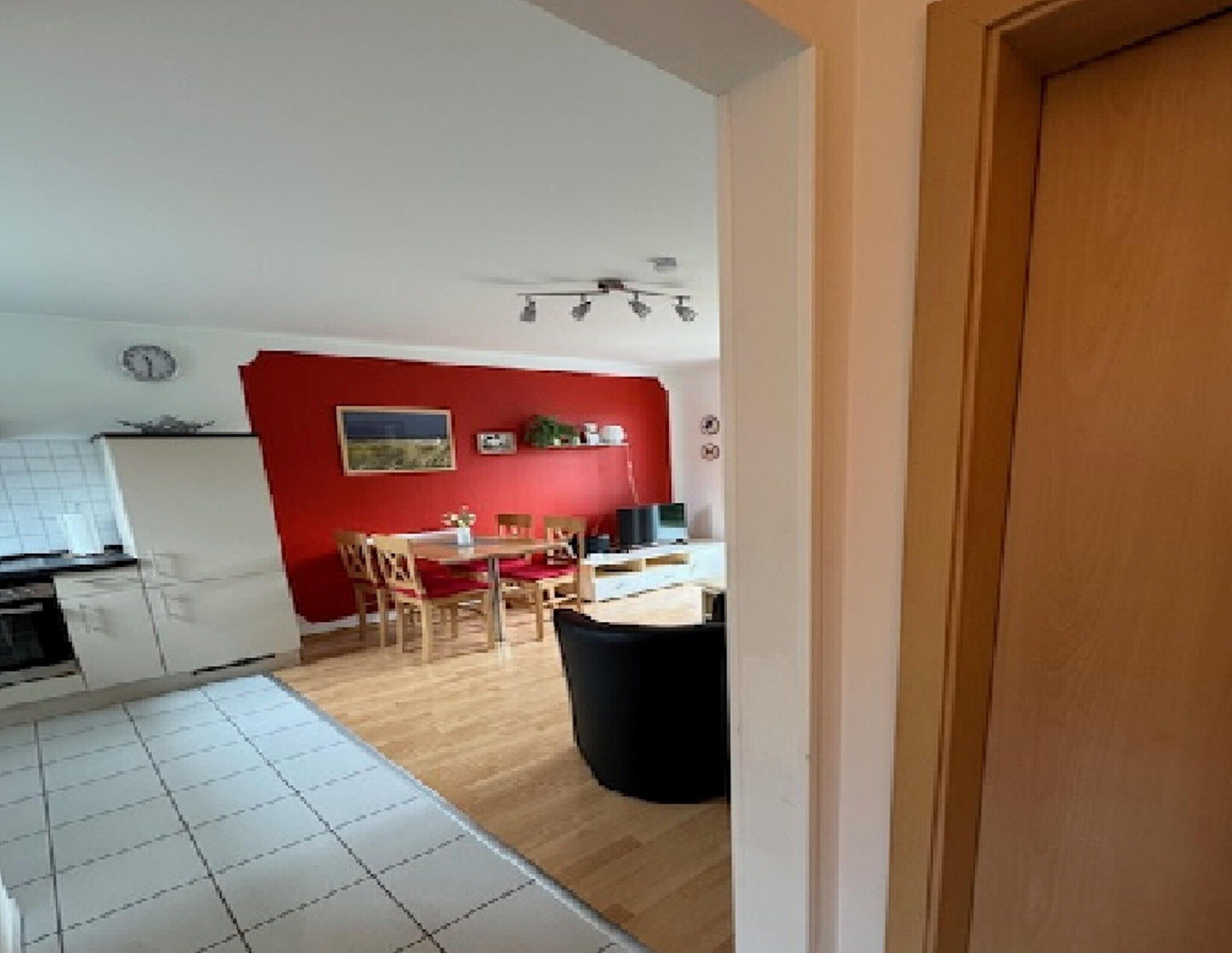 A kitchen with white cabinets, a dining area with a red wall, and a black trash bin.