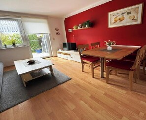 Un coin repas avec une table et des chaises en bois, un meuble TV blanc et un mur rouge orné d’une peinture représentant des fruits.