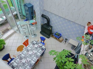 Un comedor con una mesa cubierta por un mantel estampado, rodeada de sillas. Una silla azul está al frente de la mesa, y un taburete azul se encuentra cerca de la pared. Se ve una estufa negra al fondo, y un bote de basura azul está en el suelo.