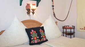 A bedroom with a wooden headboard, two white pillows, a black pillow with floral embroidery, a small wooden nightstand with a red cushion, and a wall-mounted lamp.