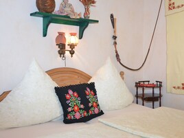 Un dormitorio con cabecera de madera, dos almohadas blancas, una almohada negra con bordado floral y una mesita de noche de madera con un cojín rojo, además de una lámpara de pared.
