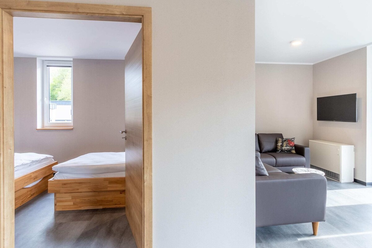 A modern, minimalist room with a wooden bed, a grey sofa, and a mounted TV.