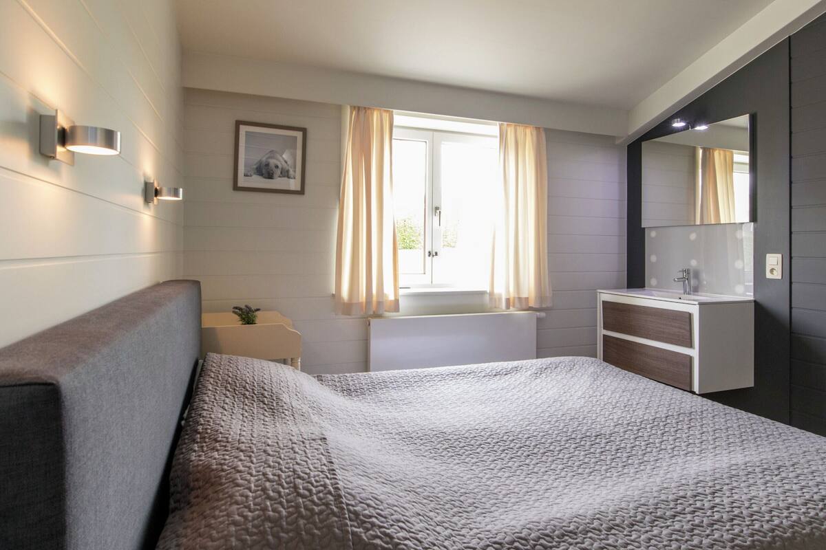 A bedroom with a bed, a bedside table, a mirror, and a window with curtains.