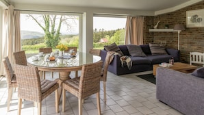 A spacious living area with a round glass table surrounded by wicker chairs, a dark-colored sofa, and a purple armchair. Large windows offer a view of greenery and hills. - Vintage Villa With Private Garden in Rendeux (Rendeux)