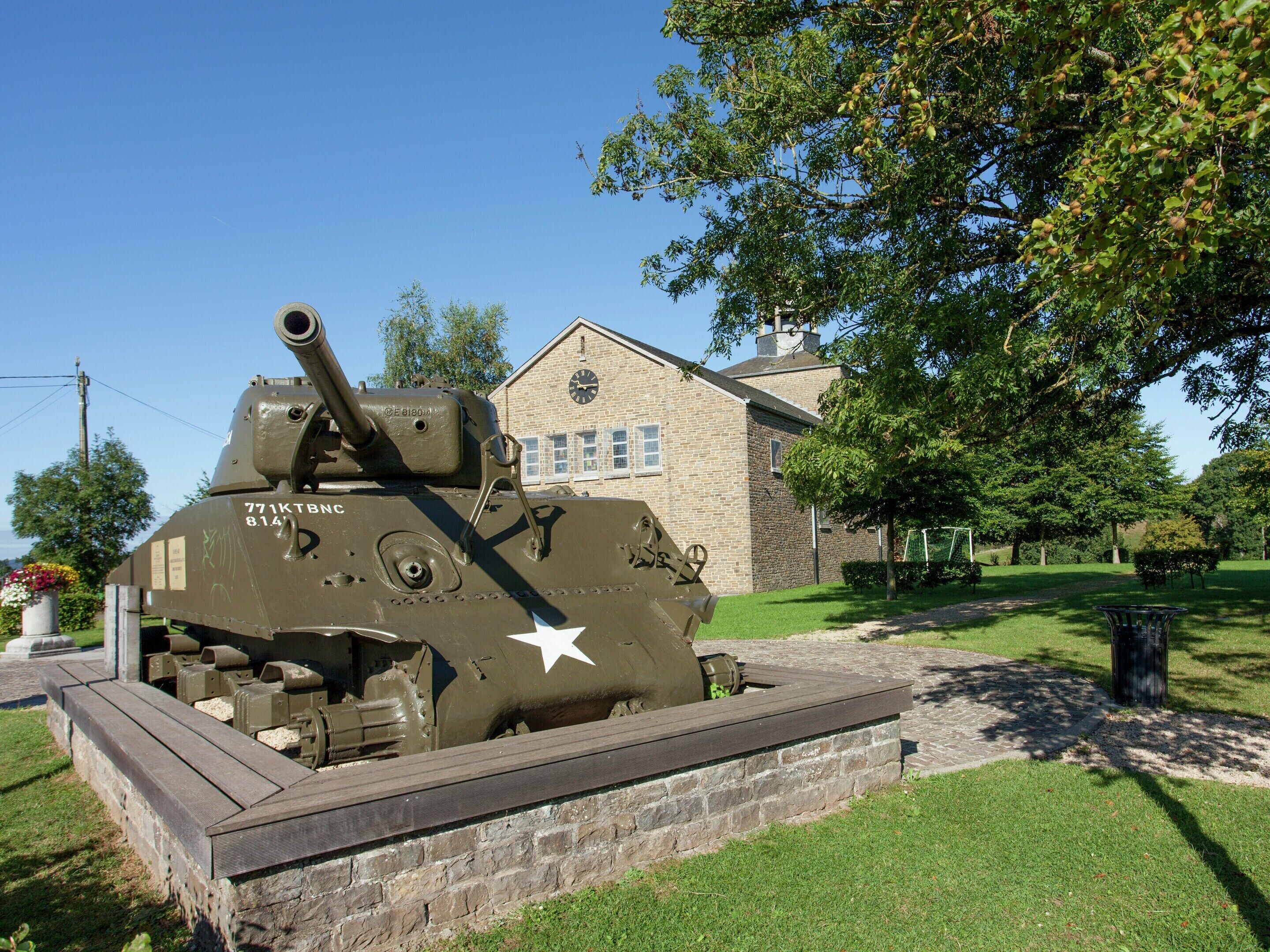 A military tank on display in a grassy area with a brick wall and a building in the background.