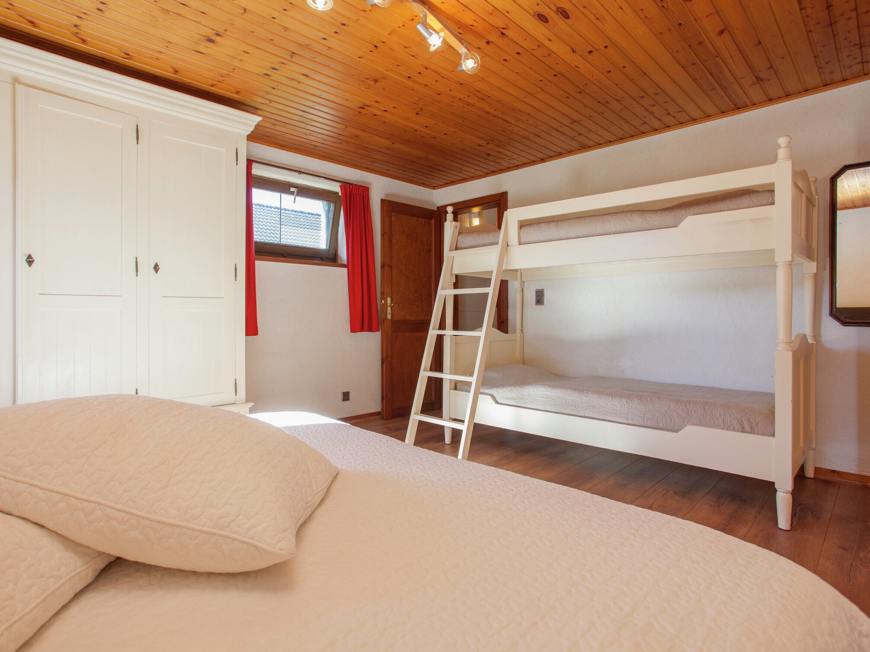 A bunk bed room with a wooden ceiling, a window with red curtains, and a white wardrobe.