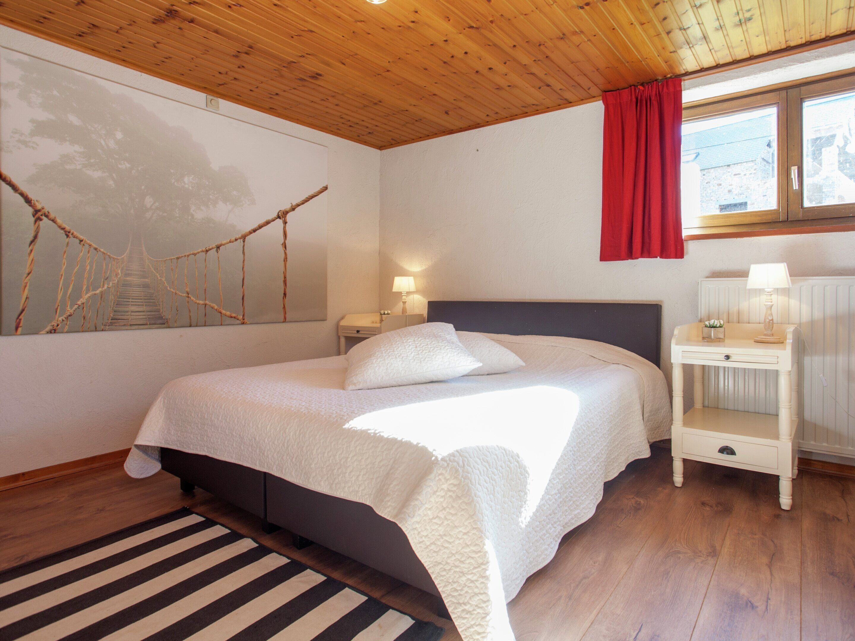 A bedroom with a bed, a nightstand, a lamp, a red curtain, and a wall painting.
