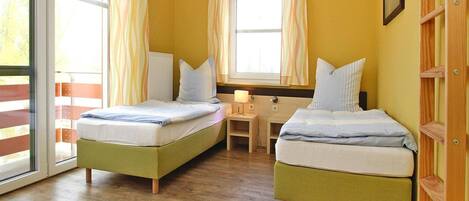 Ein Zimmer mit zwei Betten, Holzboden, einem Fenster mit Vorhängen und einem kleinen Tisch mit Lampe.