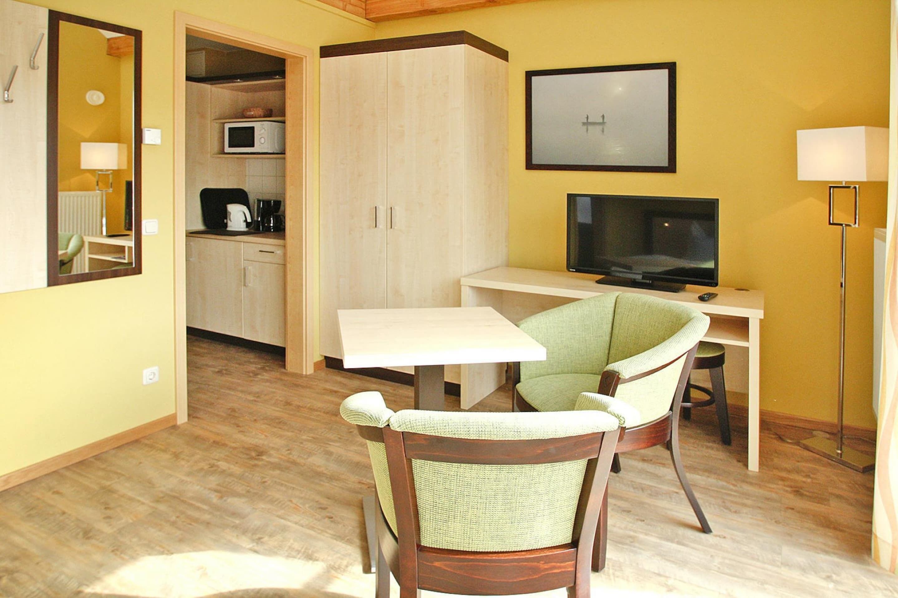 A compact room with a dining table, a green chair, a television, and a kitchen area with a microwave.