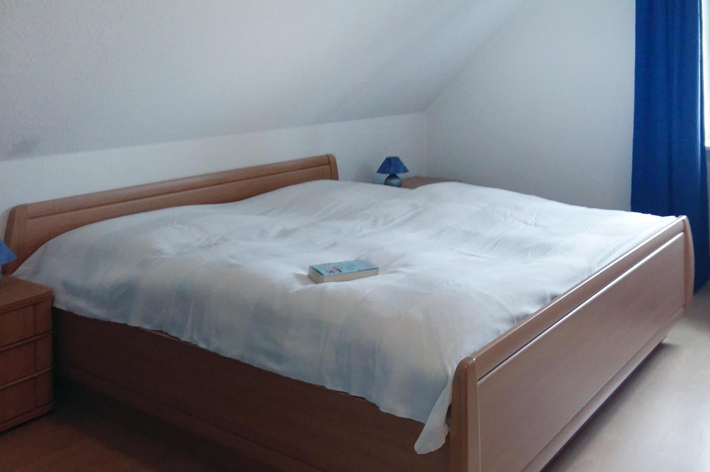 Uma cama de solteiro com cabeceira e peseira de madeira, uma mesa de cabeceira com abajur, e um livro sobre a cama.
