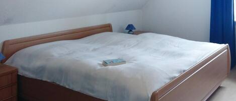 Uma cama de solteiro com cabeceira e peseira de madeira, uma mesa de cabeceira com abajur, e um livro sobre a cama.