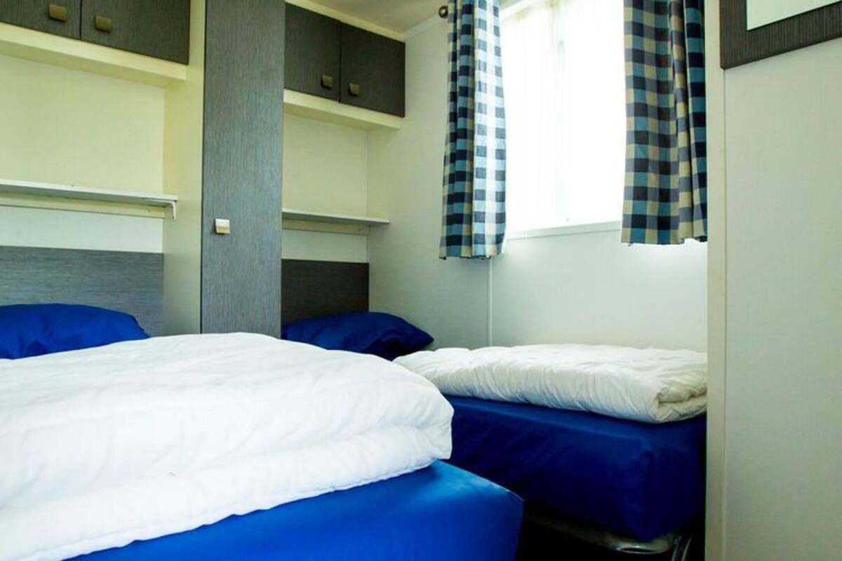 Una habitación con dos camas, cada una con colchón azul y ropa de cama blanca. Hay una ventana con cortinas a cuadros y una estantería empotrada sobre las camas.