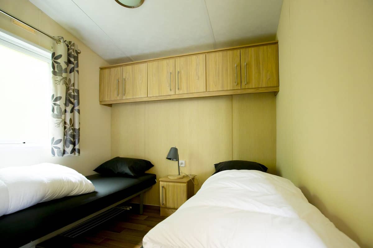 Una habitación pequeña y funcional con dos camas individuales, una mesita, una lámpara y una ventana con cortinas.