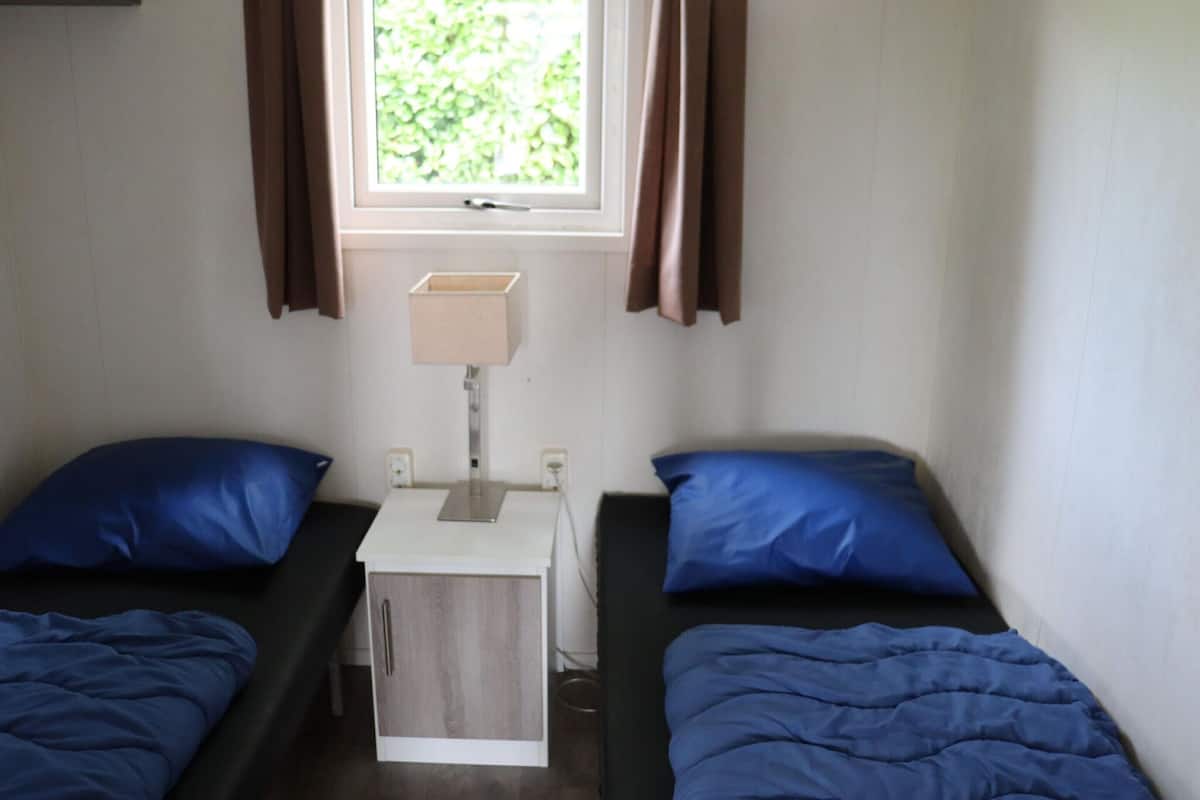 Un alojamiento pequeño y de una sola habitación con dos camas individuales, una mesita de noche con lámpara y una ventana con cortinas.