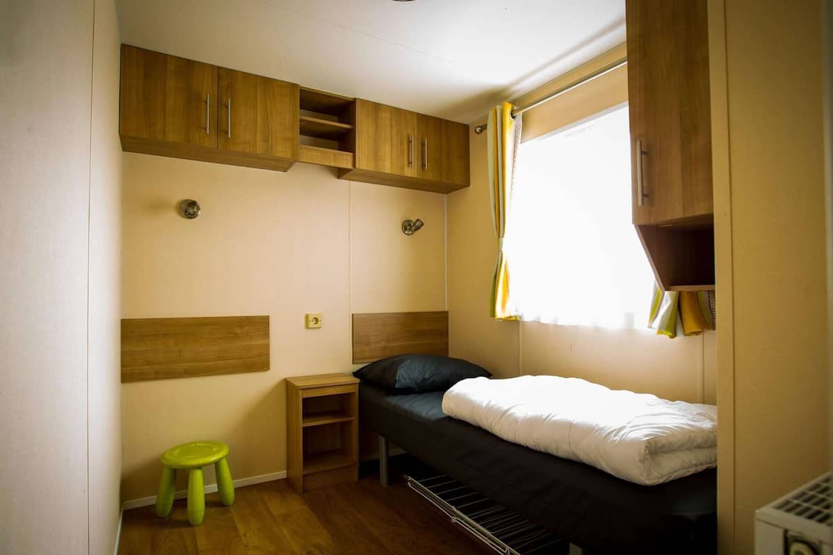 Una habitación pequeña con una litera, armarios de madera, un taburete y una ventana con cortinas.