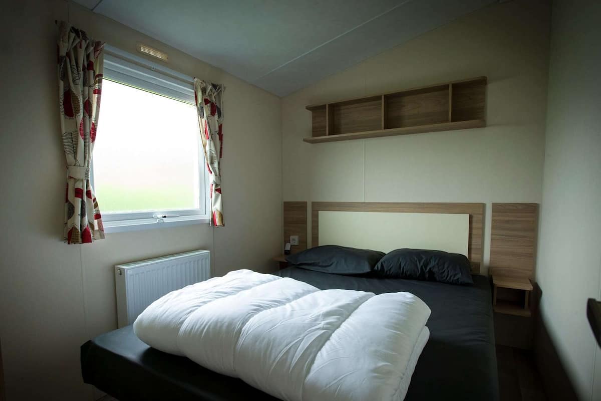 Un dormitorio con cama, ventana con cortinas, radiador y estante sobre la cama.