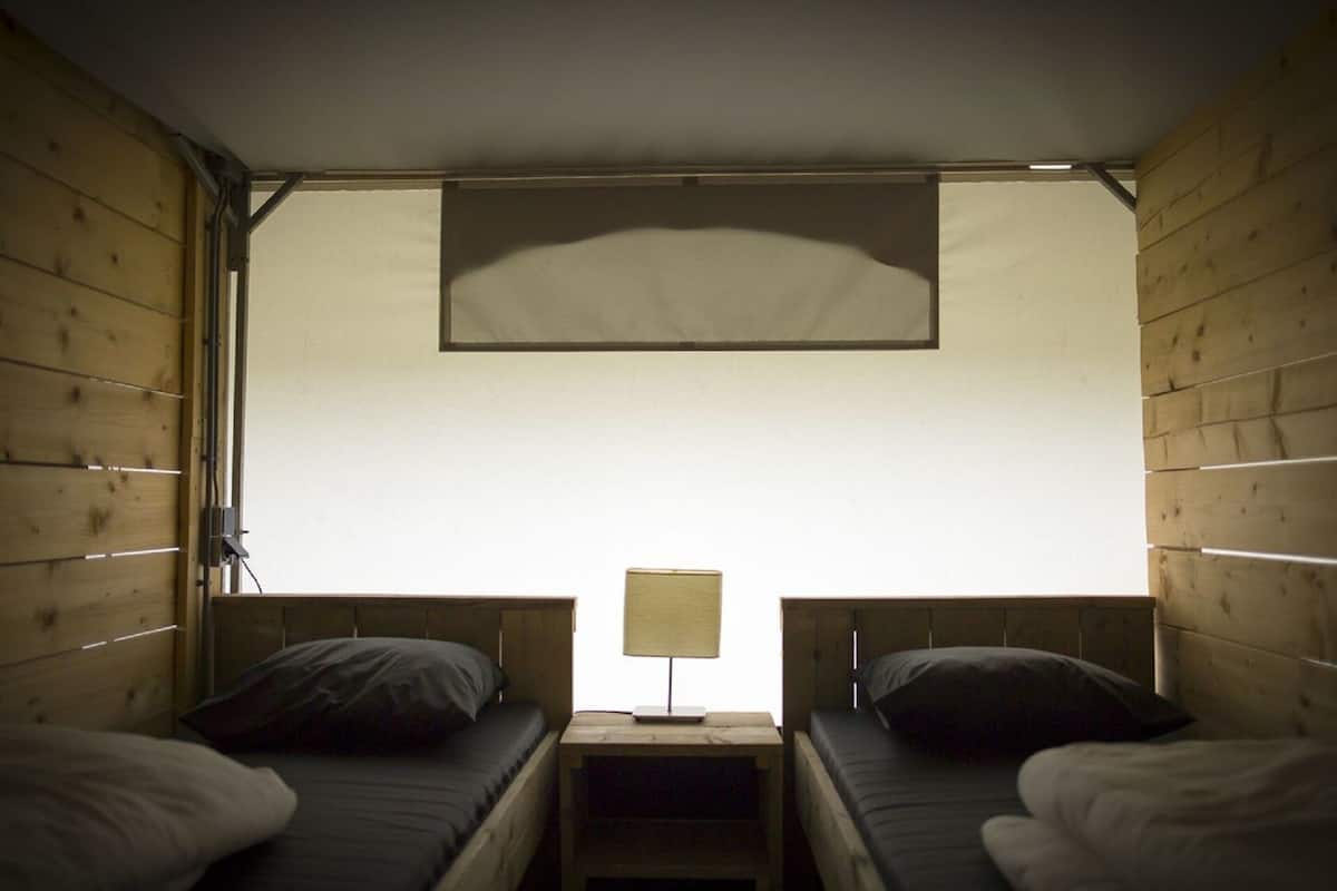 Una pequeña cabaña de una sola habitación con dos literas de madera, una mesita de noche con lámpara y un amplio ventanal que proporciona luz natural.