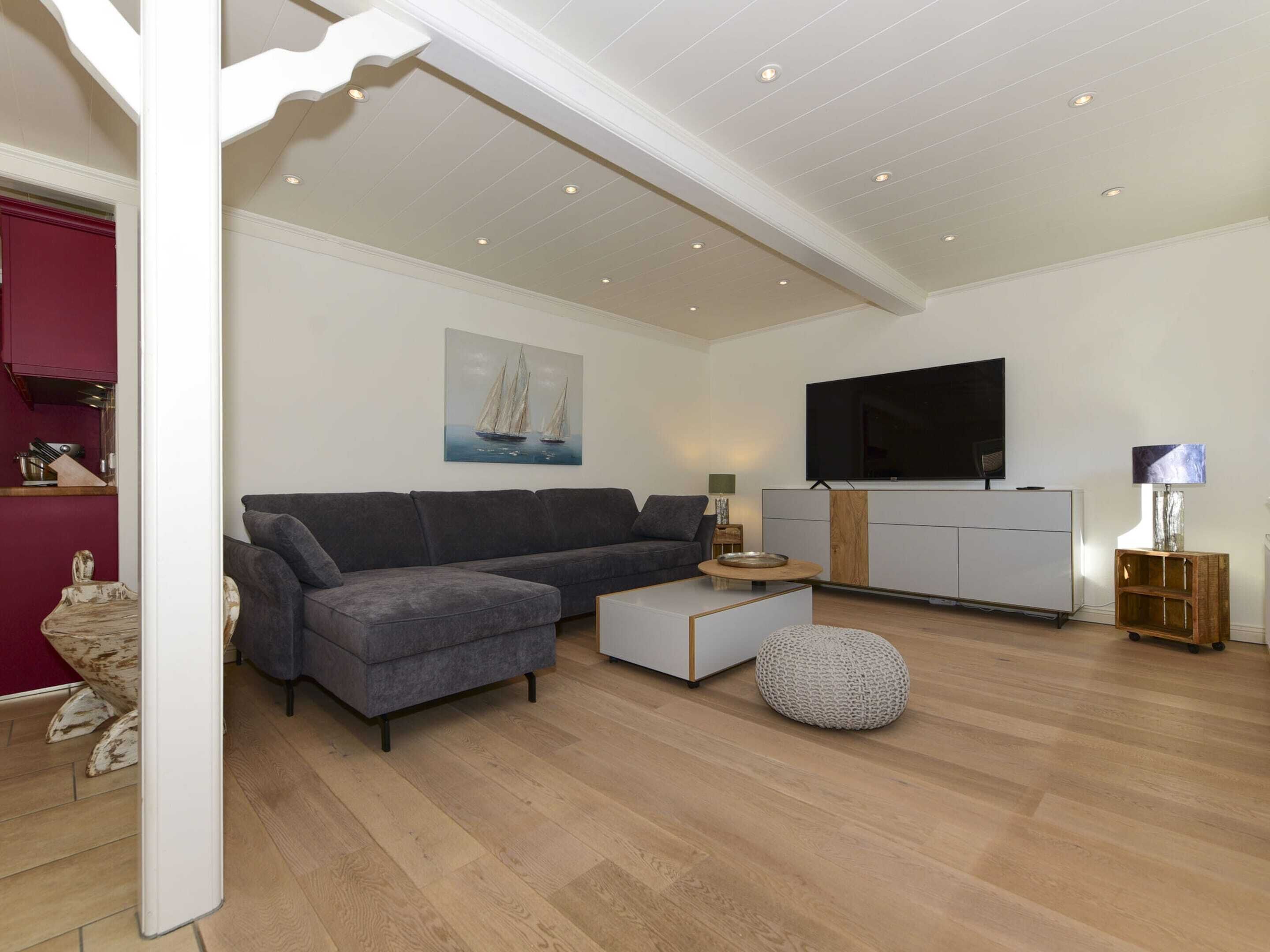 Un salon moderne avec un canapé gris, une table basse, un pouf rond, un téléviseur à écran plat et un meuble en bois.