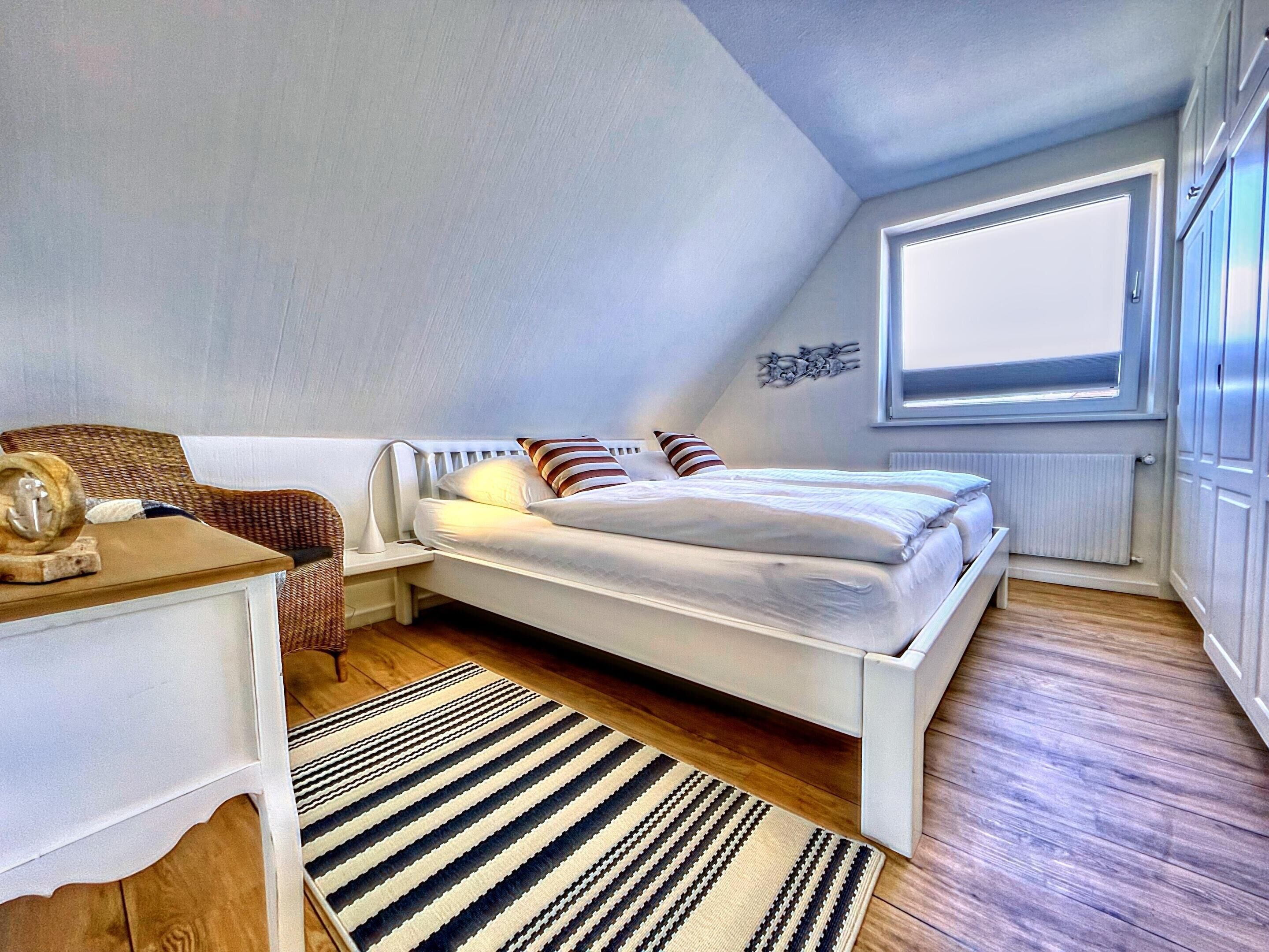 A small, well-lit bedroom with a single bed, a nightstand, a chair, and a window.