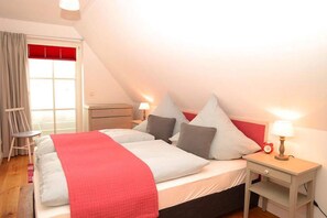 A bedroom with a bed, nightstands, a chair, a window with curtains, and a red accent wall. - Apartments, Wyk auf Foehr (Wyk auf Foehr)