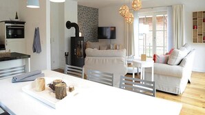 A modern kitchen with a central island, a dining area with a white sofa, and a living room with a TV and decorative lights. - Apartments, Wyk auf Foehr (Wyk auf Foehr)