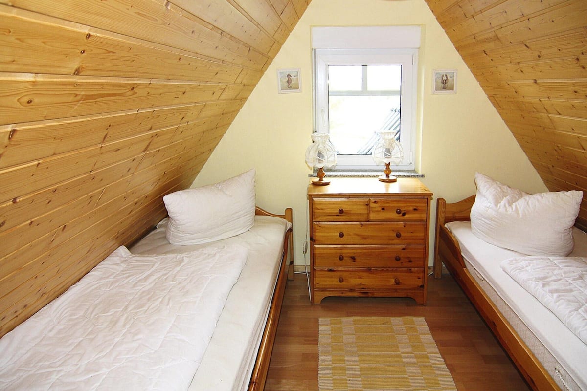 Un dormitorio acogedor en el ático, con paredes de madera y techo inclinado. Hay dos camas individuales con ropa blanca, un aparador de madera y una ventana con vistas.