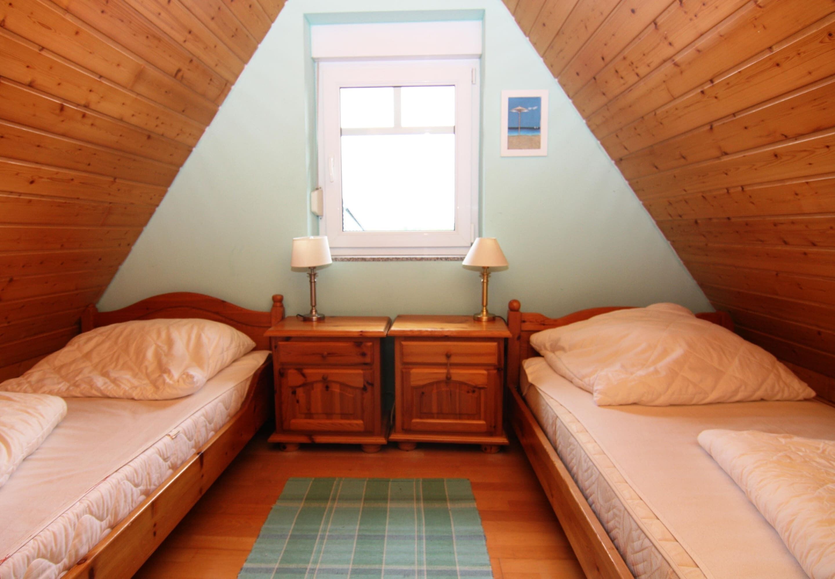 A cozy attic bedroom with two single beds, a window, and a small dresser with lamps.
