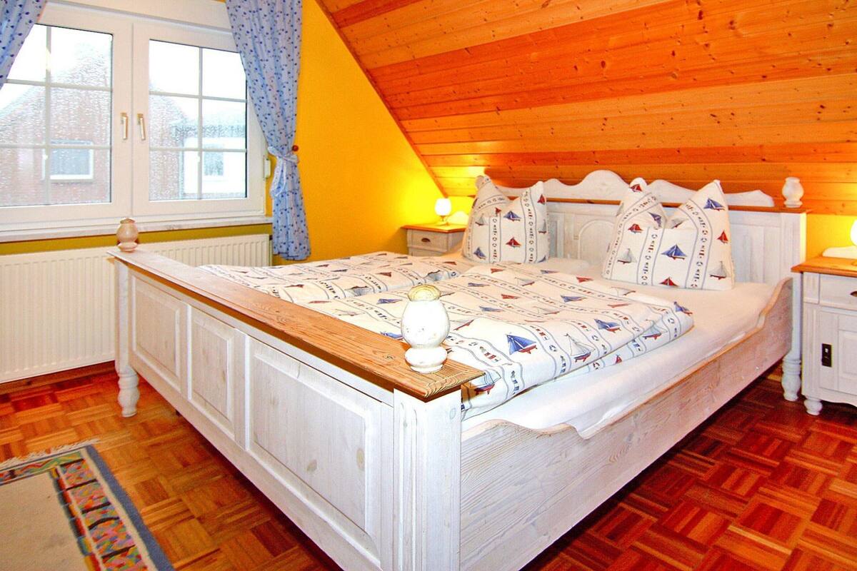 Un dormitorio con techo de madera, una cama con cabecera blanca, una mesita de noche con lámpara y una ventana con cortinas.