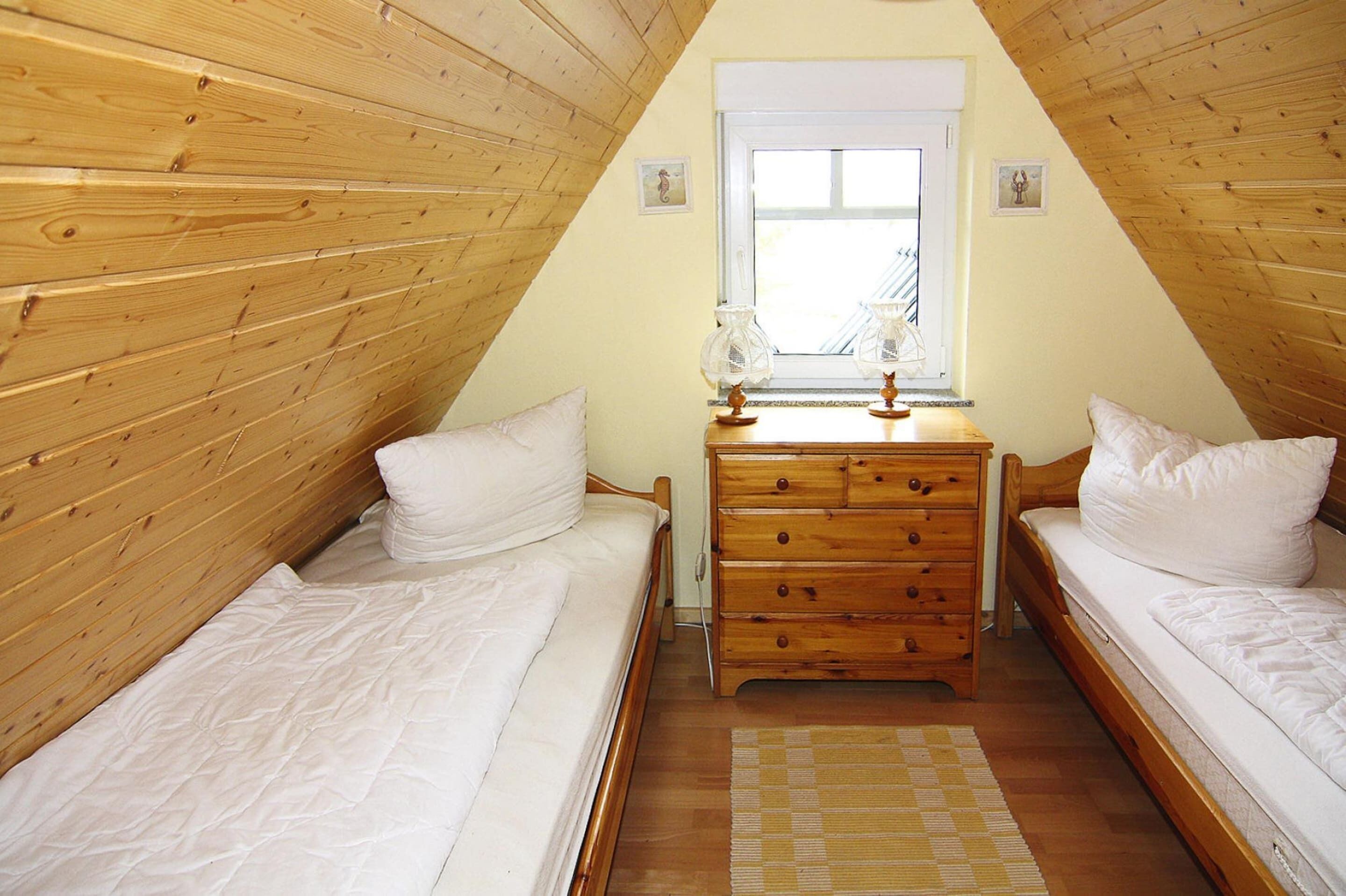 A small, cozy attic bedroom with wooden walls and a sloped ceiling. There are two single beds with white bedding, a wooden dresser with drawers, and a window with a view of the outside.