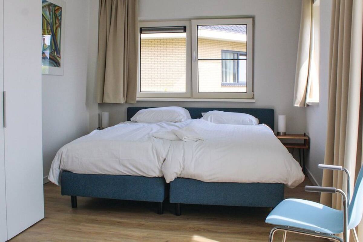 Un lit double avec une tête de lit, une table de chevet, une chaise et une fenêtre avec des rideaux.