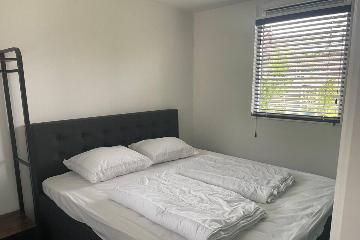 Un lit simple avec une tête de lit sombre, du linge de lit blanc et une fenêtre avec des stores.