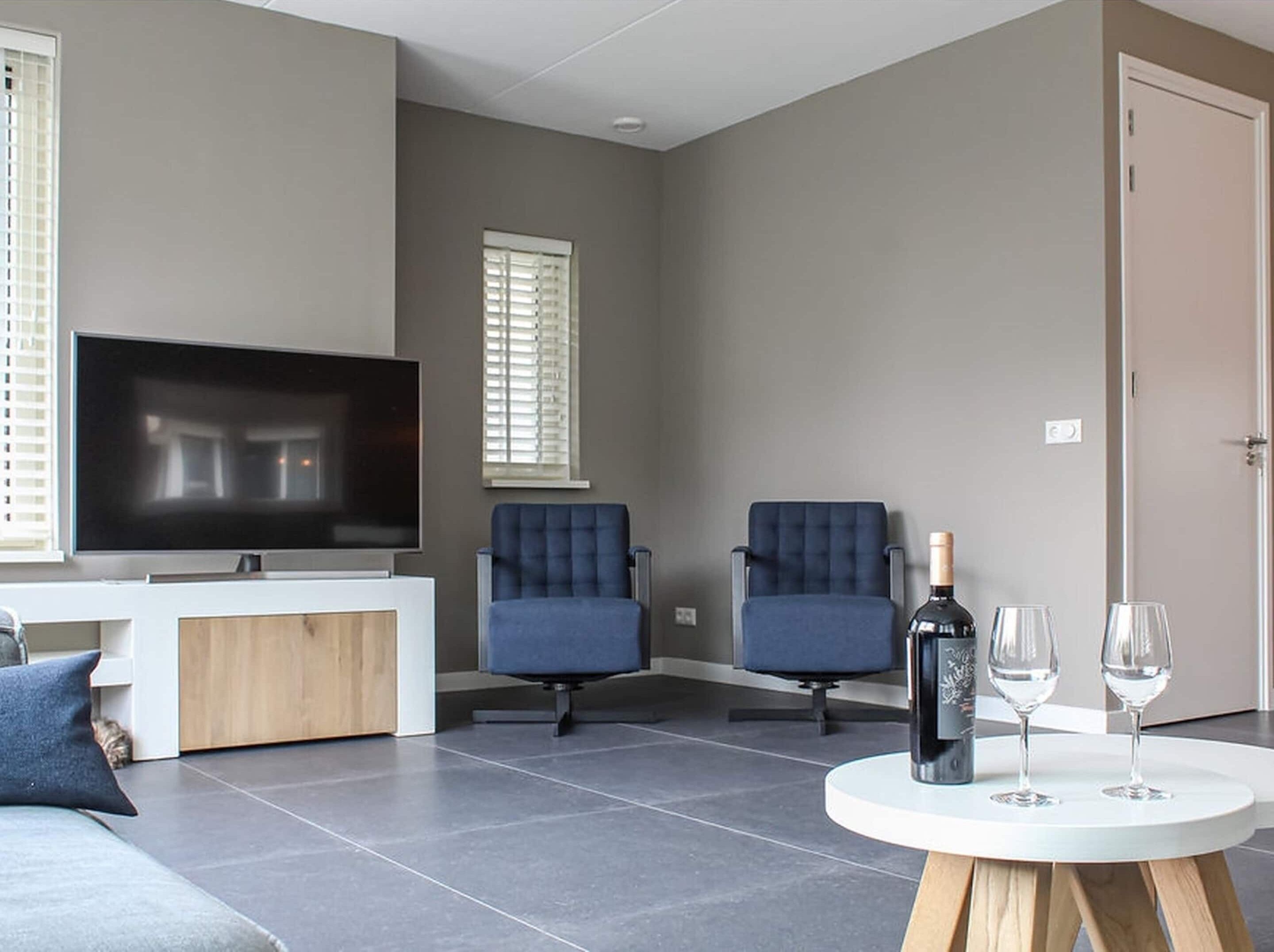 Un salon moderne doté d’un téléviseur à écran plat, de deux fauteuils bleus, d’une table basse en bois avec deux verres à vin et d’une porte.
