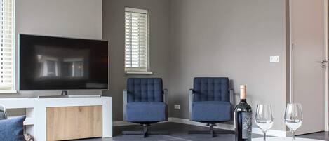 Un salon moderne doté d’un téléviseur à écran plat, de deux fauteuils bleus, d’une table basse en bois avec deux verres à vin et d’une porte.