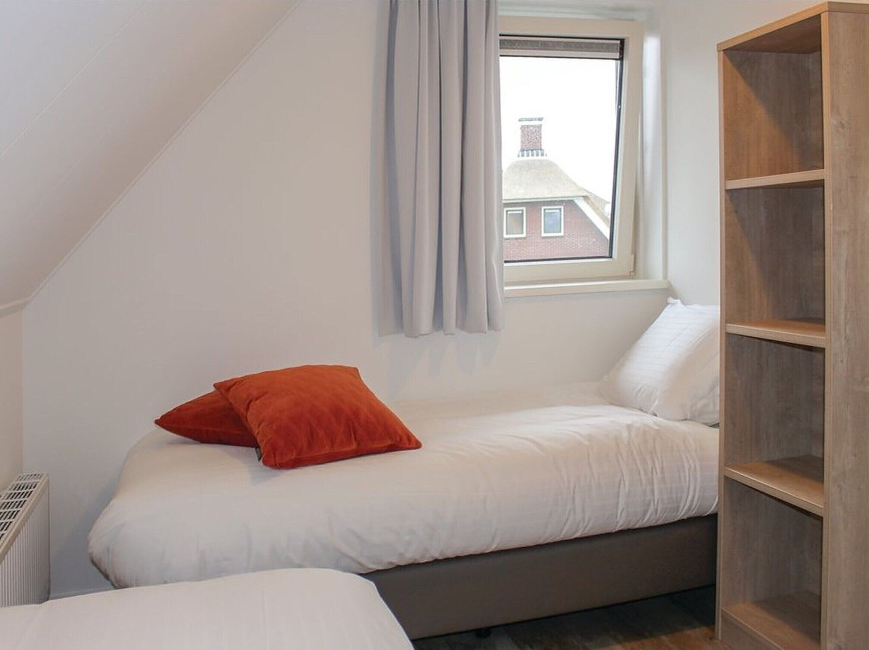 Una habitación pequeña con una cama individual, techo inclinado, ventana con cortinas y estantería de madera.