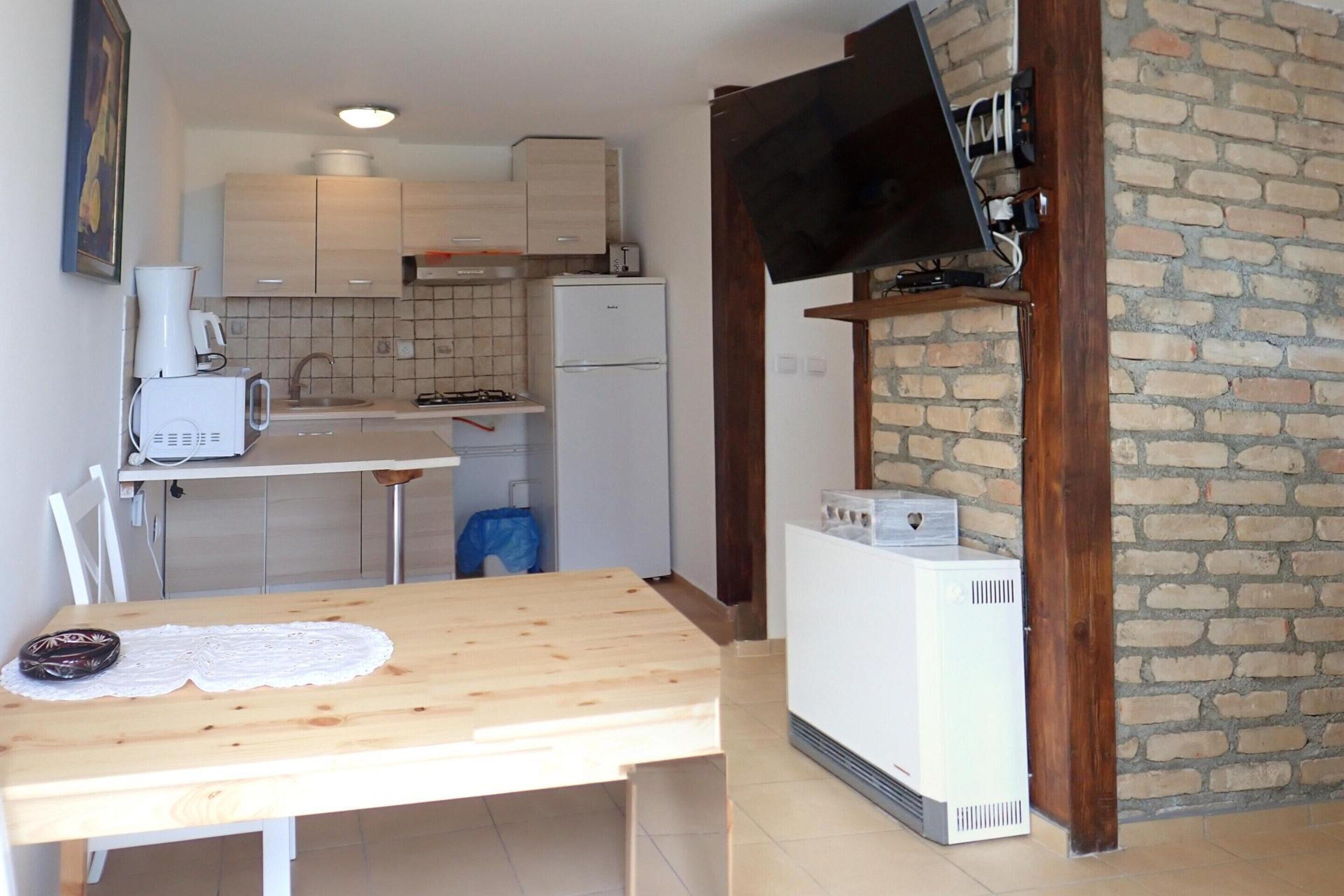 A compact kitchen with a wooden table, a white refrigerator, and a TV mounted on a brick wall.