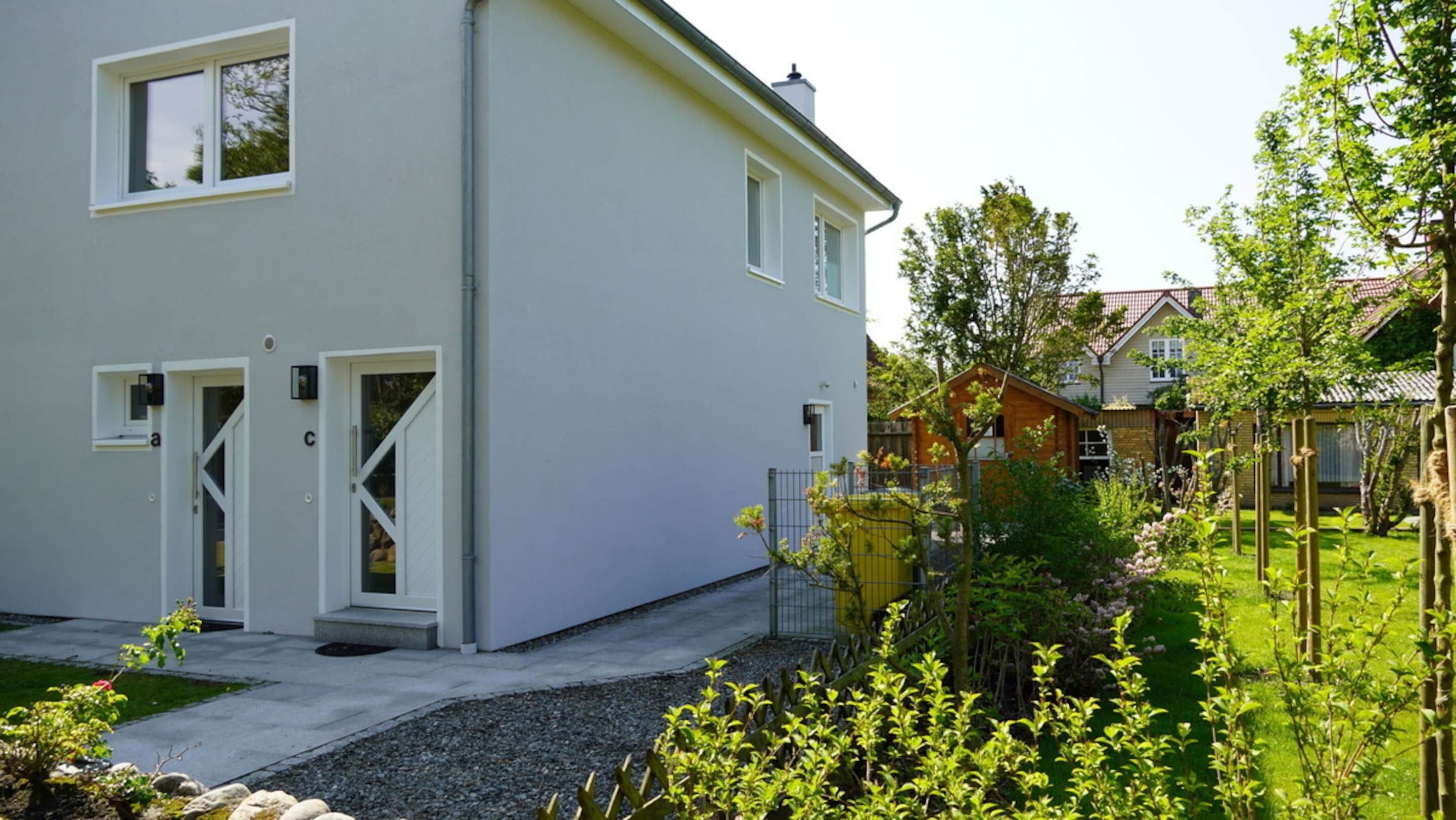 Ein modernes, zweistöckiges Haus mit weißer Fassade, einer Haustür und einem kleinen Garten mit Zaun und Bäumen.