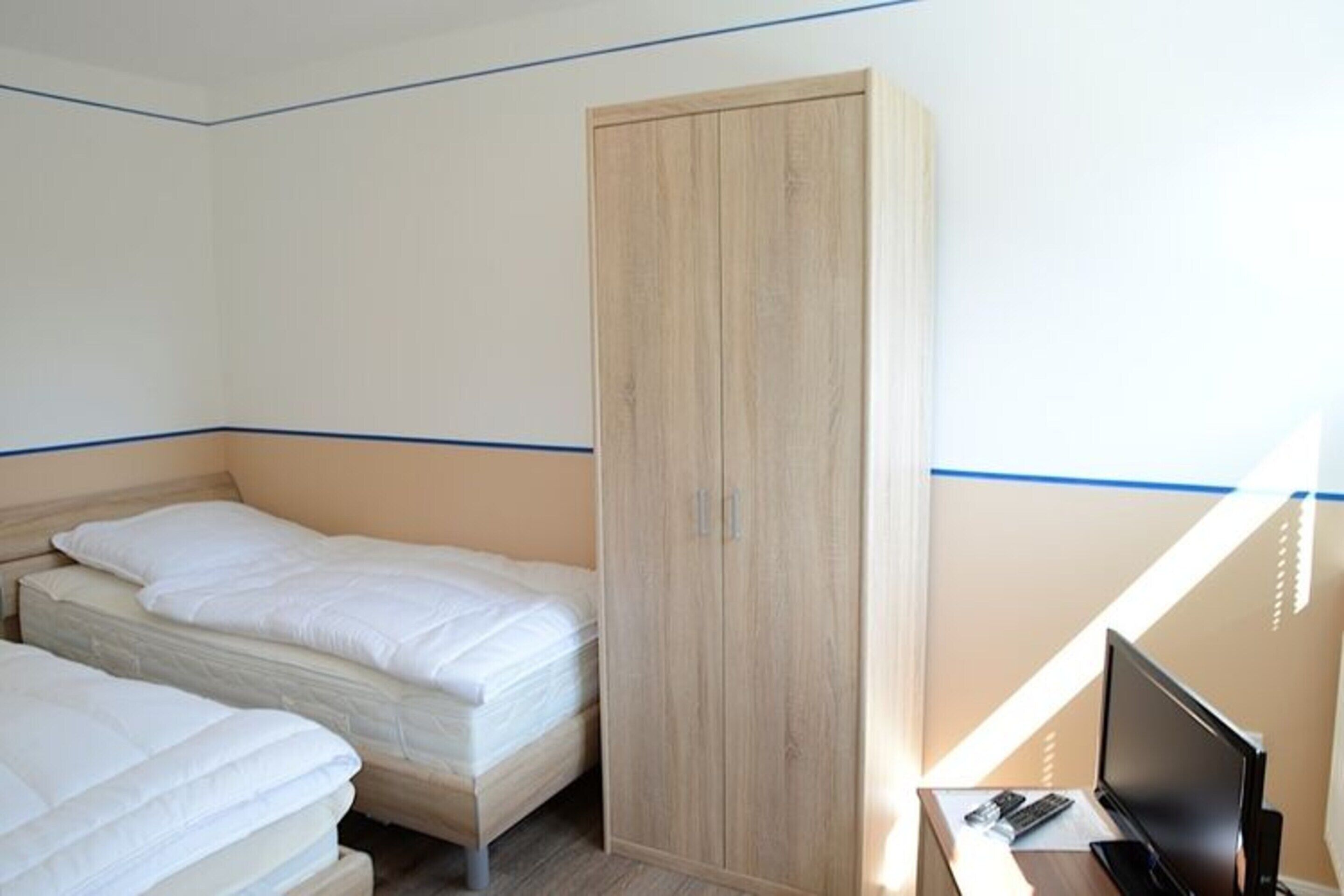 A hotel room with two single beds, a wooden wardrobe, a desk with a computer, and a TV.