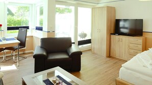 A modern room with a sofa, a TV, a dining area with a table and chairs, and a large window offering a view of greenery.