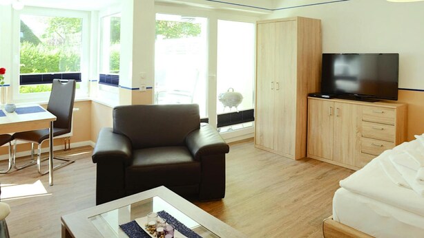 A modern room with a sofa, a TV, a dining area with a table and chairs, and a large window offering a view of greenery. - Apartment House, Wyk auf Foehr (Wyk auf Foehr)