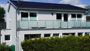 A modern two-story building with a balcony on the upper floor, a satellite dish on the roof, and a green hose visible on the ground.