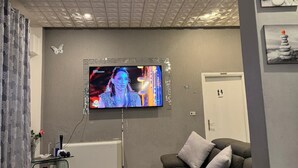 Een woonkamer met een flatscreen televisie aan de muur, een grijze bank, een kleine tafel met een vaas en een decoratieve vlinder op de muur.