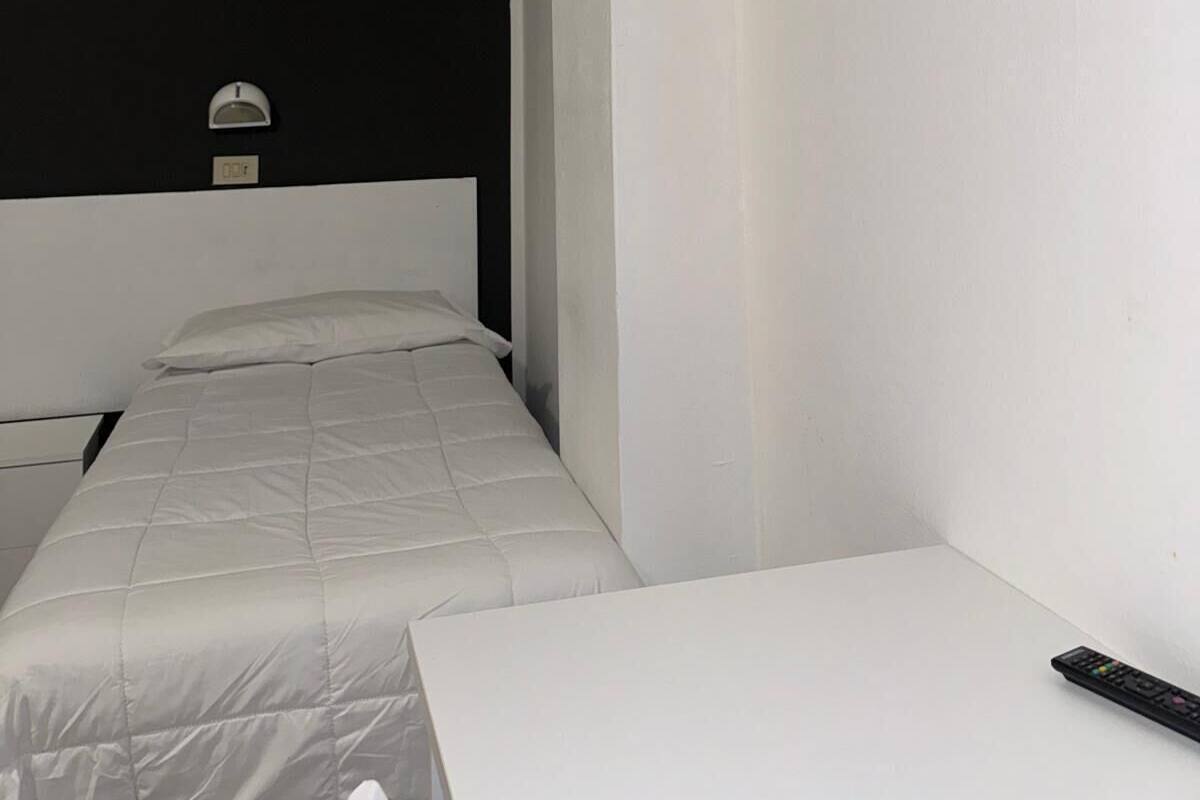 Een kleine kamer met een enkel bed, een wit hoofdbord, nachtkastje en een schilderij van een strandgezicht aan de muur.