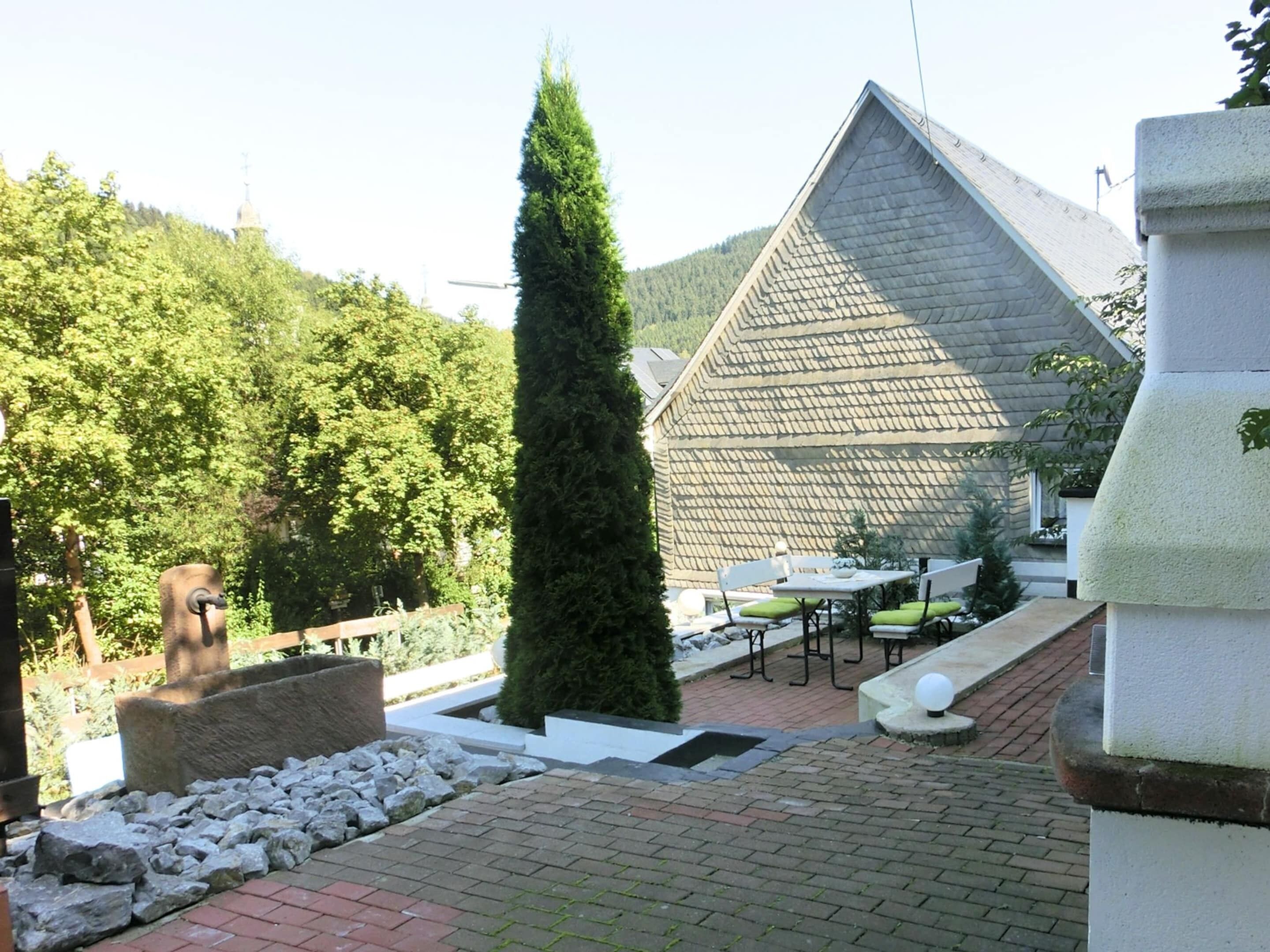 A courtyard with a stone bench, a tall coniferous tree, and a building with a sloped roof.