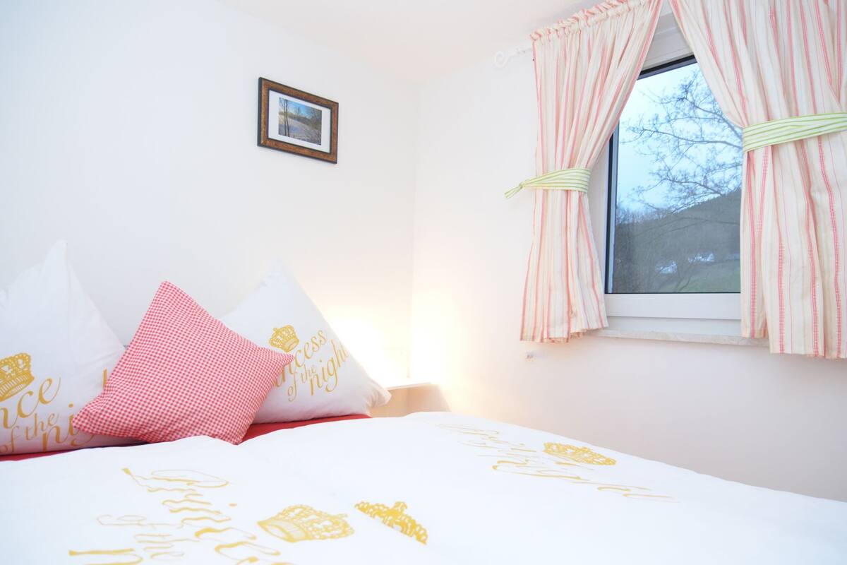 Een slaapkamer met een bed, kussens, een lijst met een afbeelding aan de muur en een raam met gordijnen.