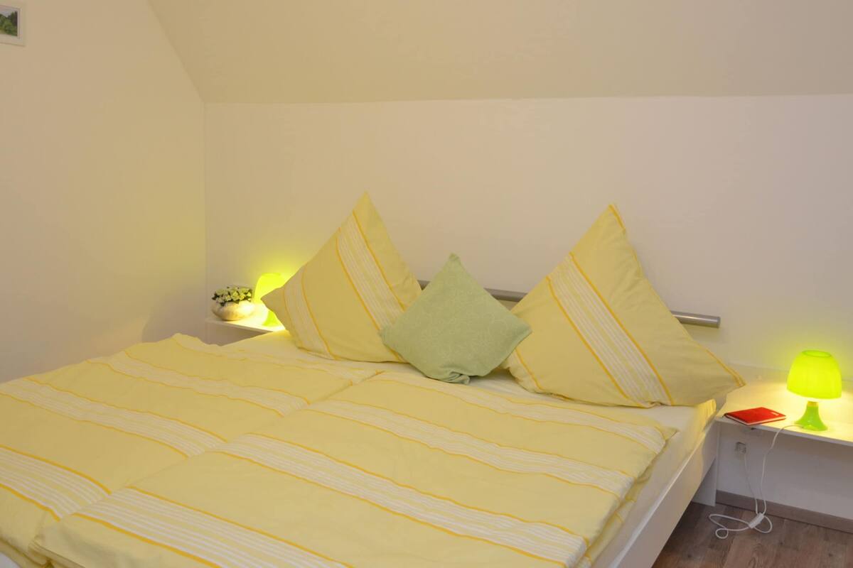 Een opgemaakt bed met gele beddengoed en kussens, een nachtkastje met een lamp en een kleine plant erop.